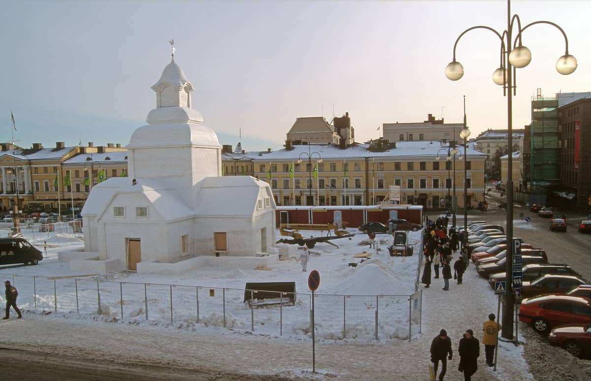 Praça da cidade coberta de neve em Helsinki com uma grande escultura de neve em forma de edifício, pessoas caminhando e carros estacionados ao longo da rua em um frio dia de inverno.