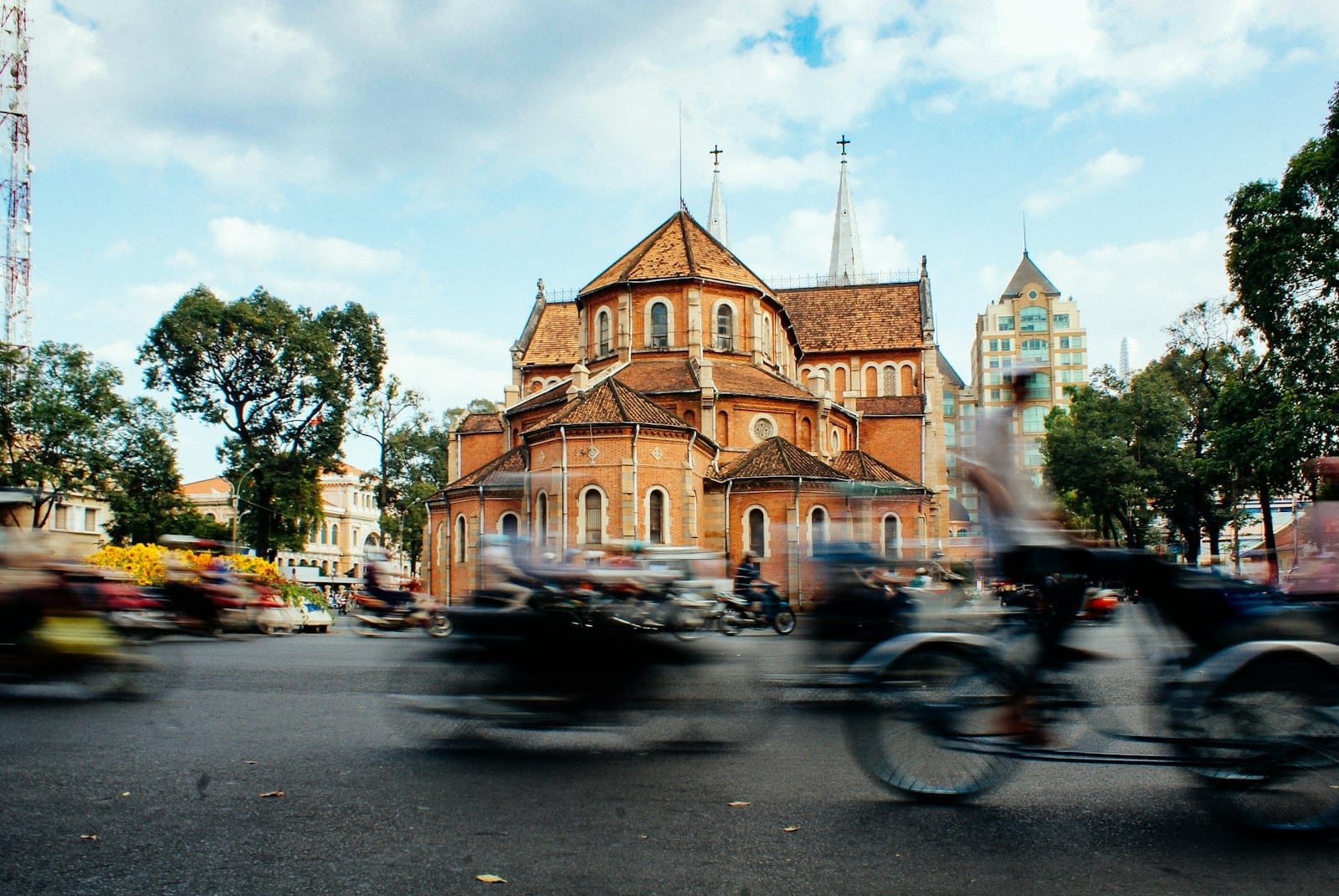 Motocicletas pasando frente a la Basílica Catedral de Notre-Dame de Saigón bajo un cielo azul con algunas nubes en Ciudad Ho Chi Minh.