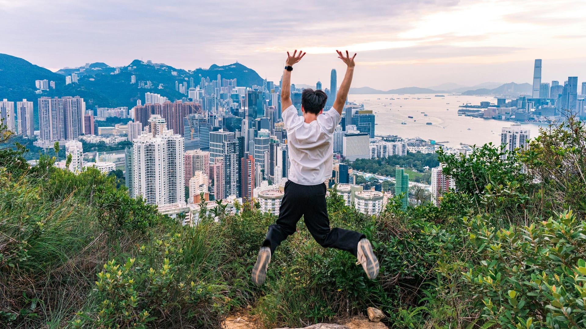 Pessoa pulando em uma colina com vista para o skyline de Hong Kong e Victoria Harbour