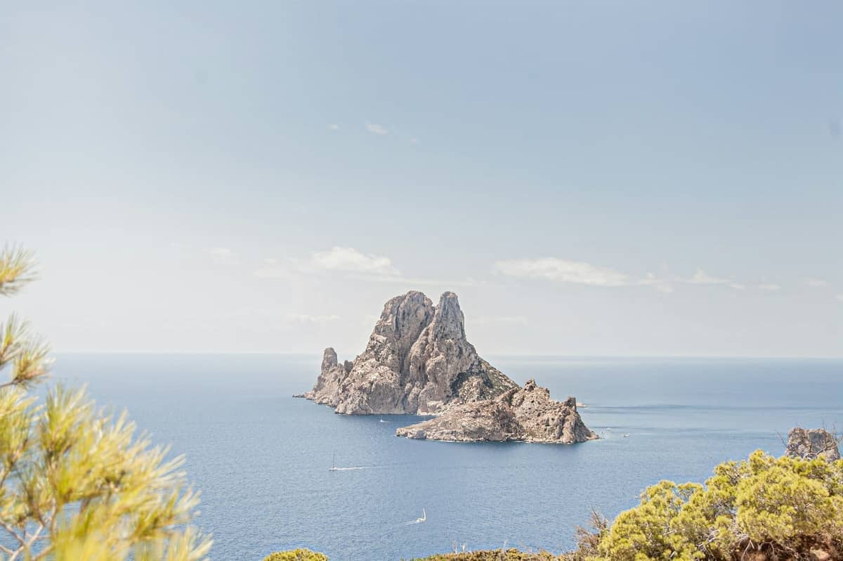 Bright, clear view of Es Vedrà island off Ibiza, with vivid blue sky, calm sea, and sunlit Mediterranean vegetation in the foreground.