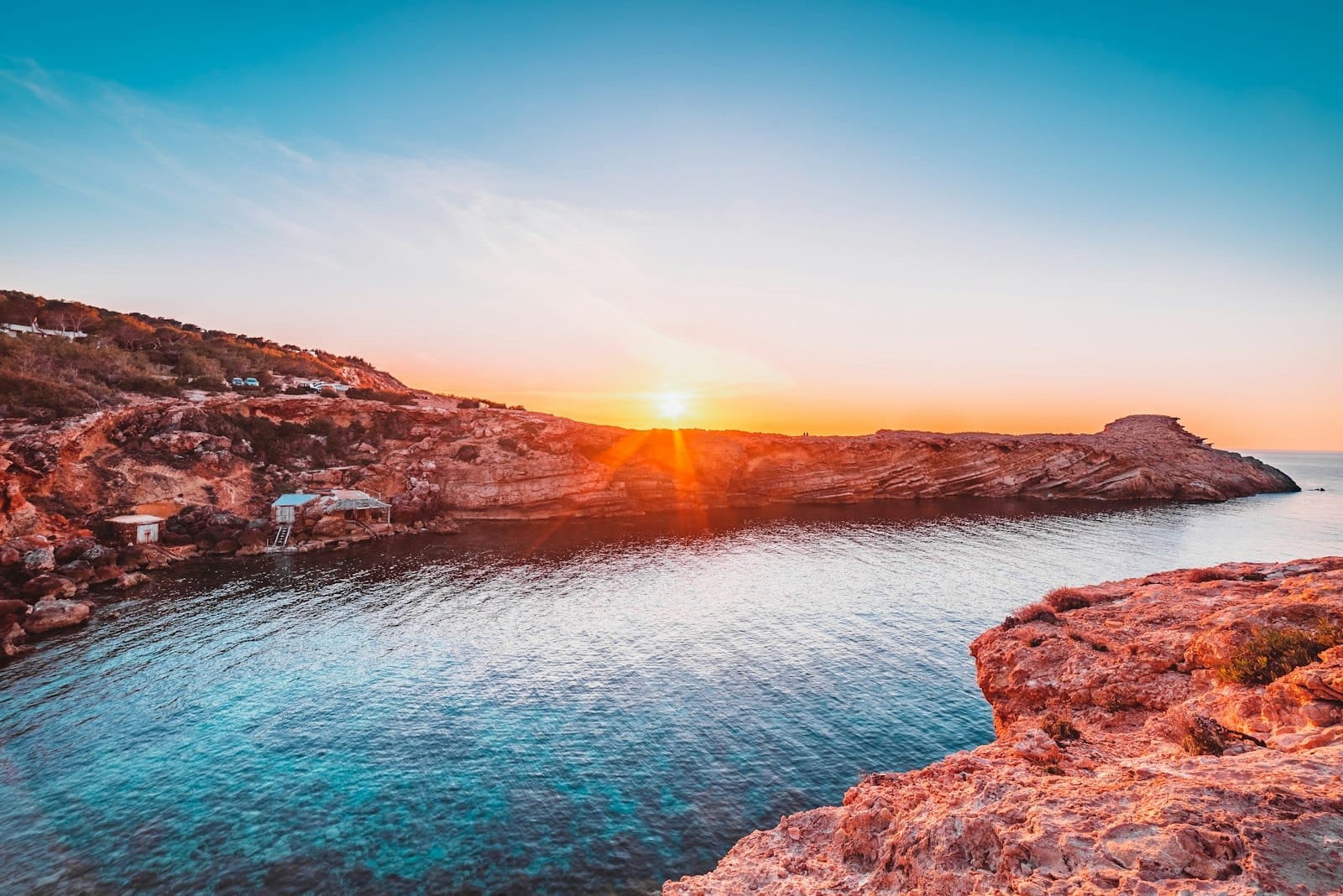 Costa rochosa e água turquesa na praia Cala Comte de Ibiza, com o sol se pondo atrás de falésias escarpadas sob um céu limpo.