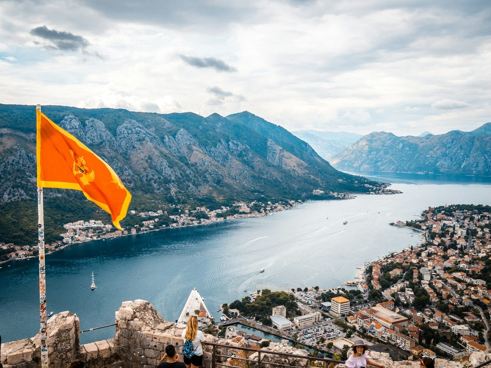 Vista panorâmica da Baía de Kotor com montanhas, edifícios dispersos e a bandeira montenegrina em primeiro plano sob um céu nublado.