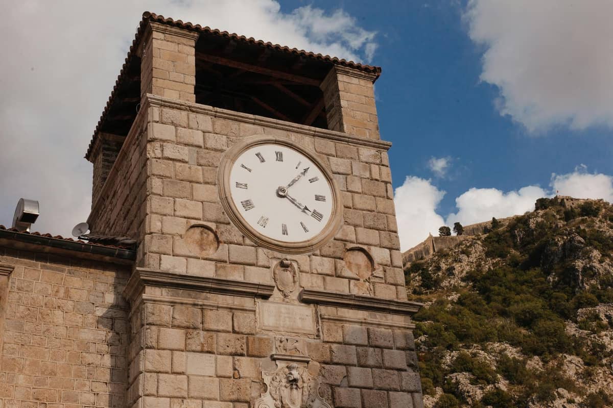 Stone clock tower in Kotor Old Town with Roman numerals and blue sky, with mountains and fortress visible in the background.