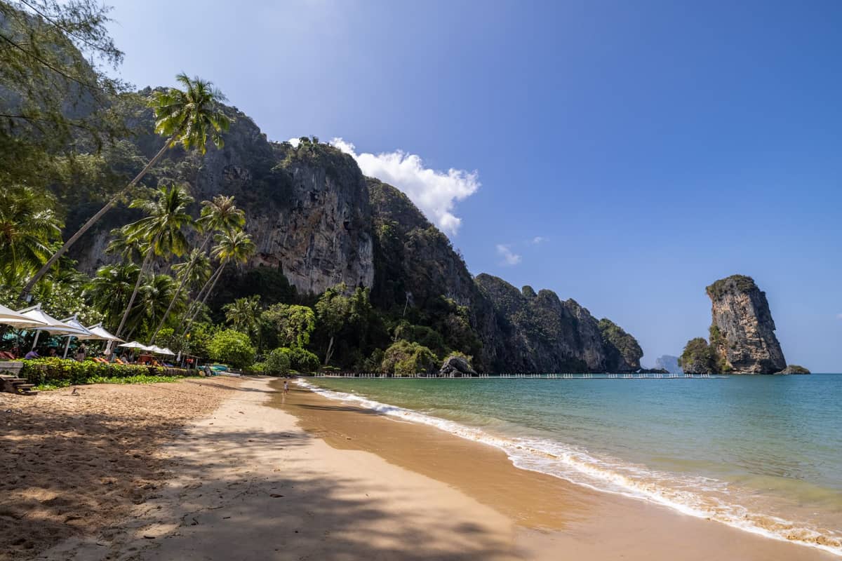 Słoneczna tropikalna plaża w Krabi z klifami wapiennymi, krystalicznie czystą wodą, palmami i pustymi leżakami wzdłuż piasku pod błękitnym niebem.