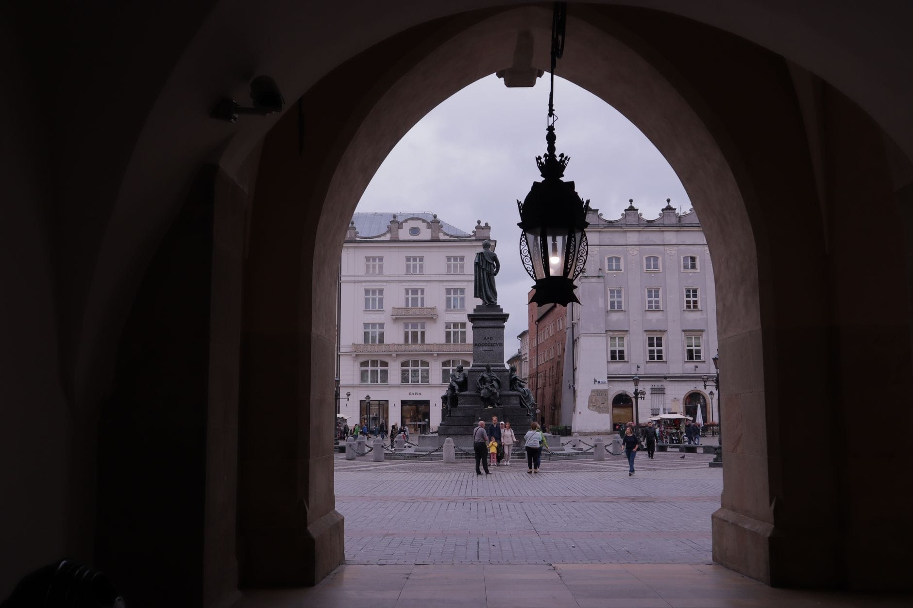 วิวผ่านซุ้มหินสู่จัตุรัสหลักของเมือง Kraków ที่มีอนุสาวรีย์ Adam Mickiewicz และอาคารสีขาวทางประวัติศาสตร์ภายใต้ท้องฟ้าครึ้ม