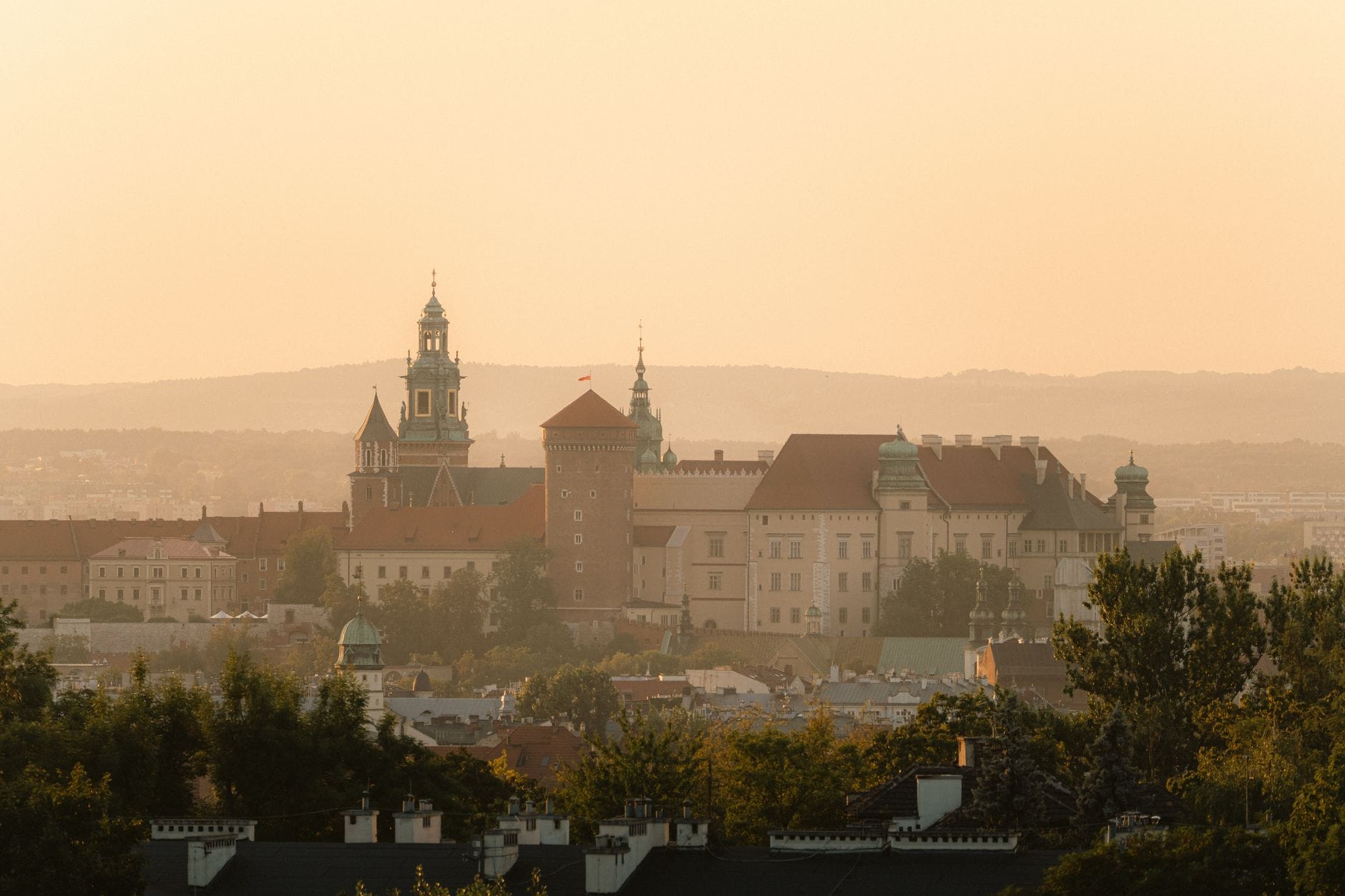 ปราสาท Wawel ในเมือง Kraków ที่ถูกโอบล้อมด้วยแสงสีทองอบอุ่นของฤดูใบไม้ร่วง มีต้นไม้อยู่เบื้องหน้าและเนินเขาอ่อนโยนเป็นฉากหลัง