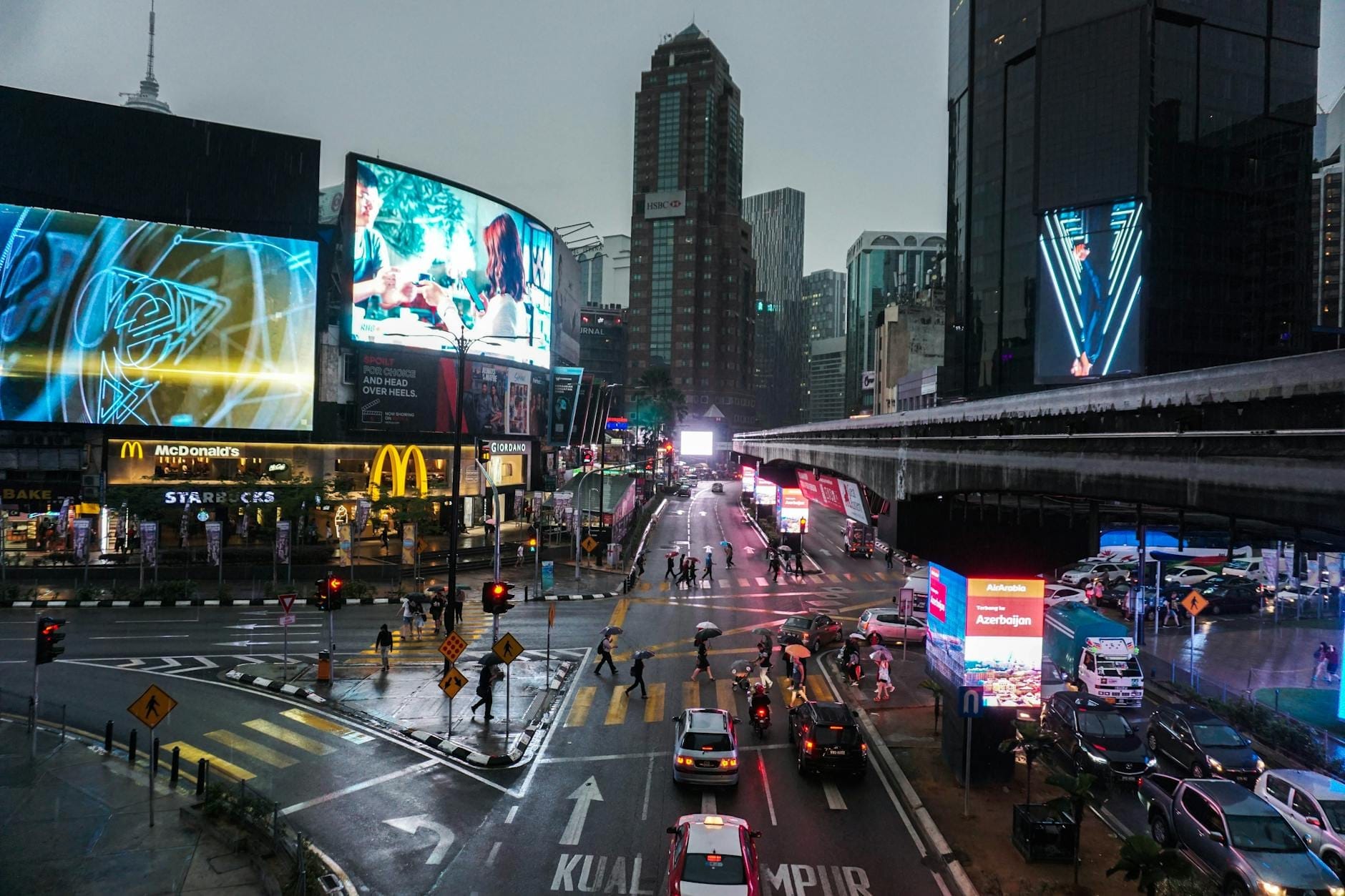 Rainy evening at Bukit Bintang intersection in Kuala Lumpur, with illuminated billboards, busy traffic, pedestrians, and iconic fast food signs.