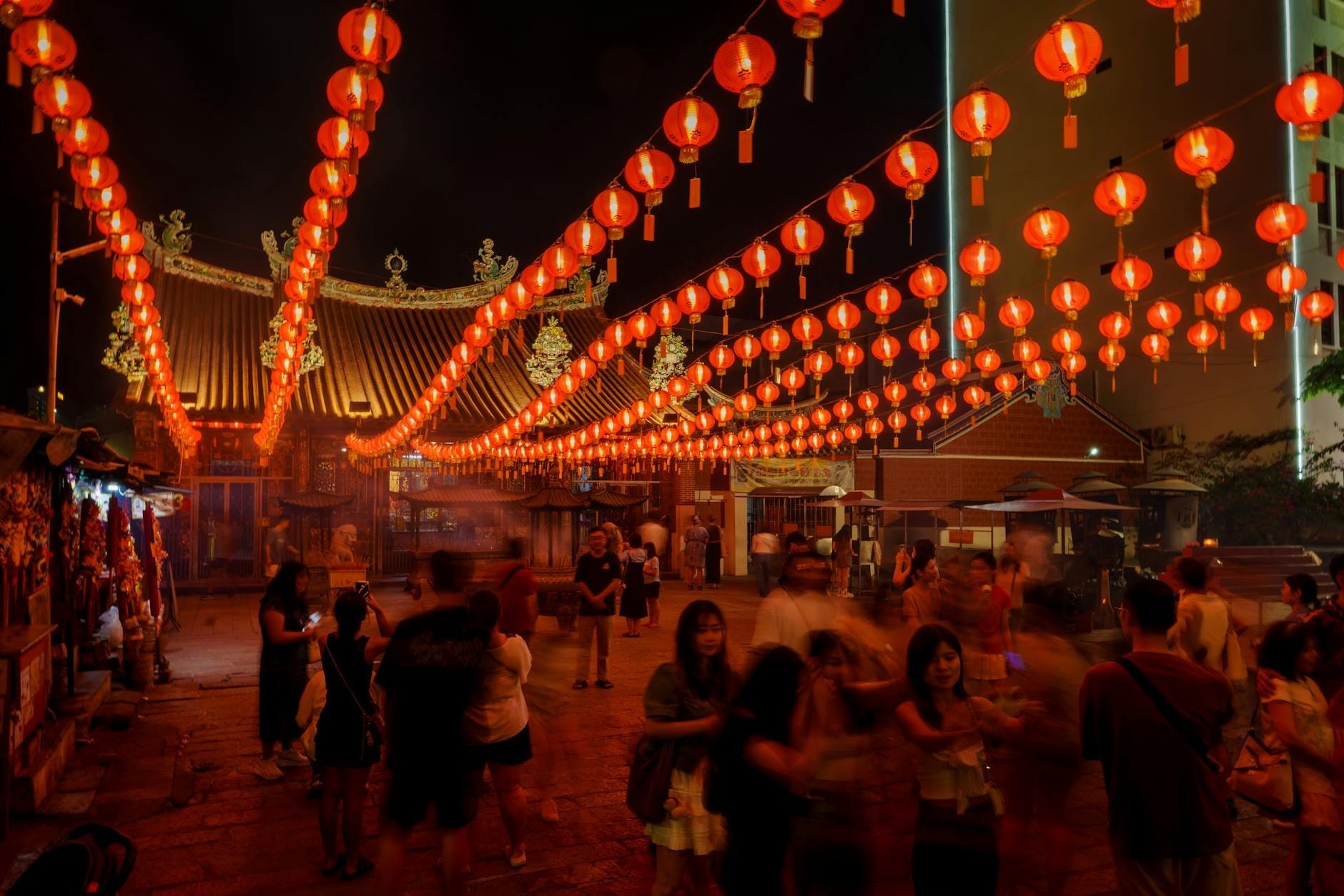 Warm red lantern lights cast a magical glow over a courtyard, creating a lively and celebratory nighttime atmosphere in Kuala Lumpur during Chinese New Year festivities