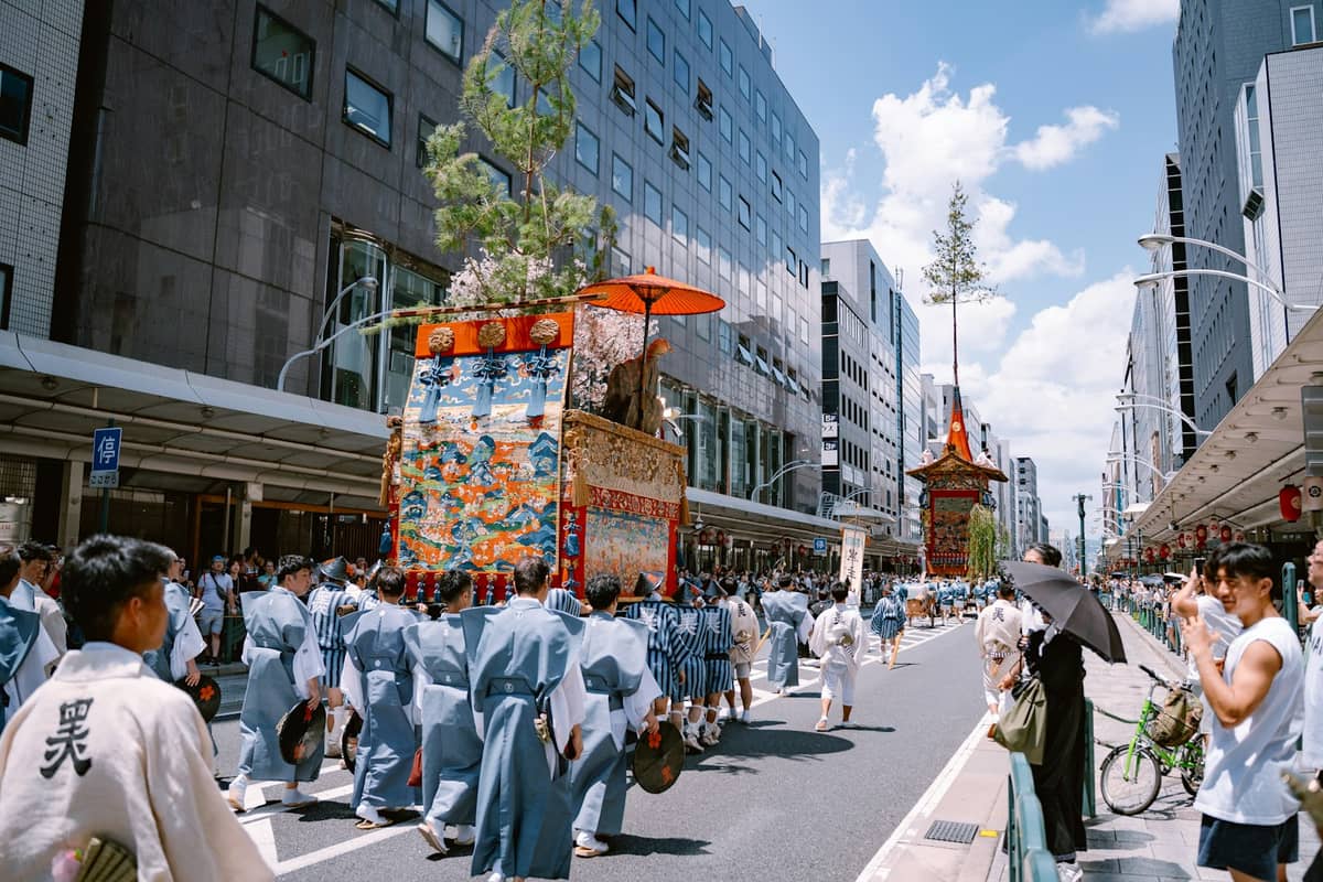 Large parade float and people in traditional attire participating in a summer festival on a sunny street in Kyoto.