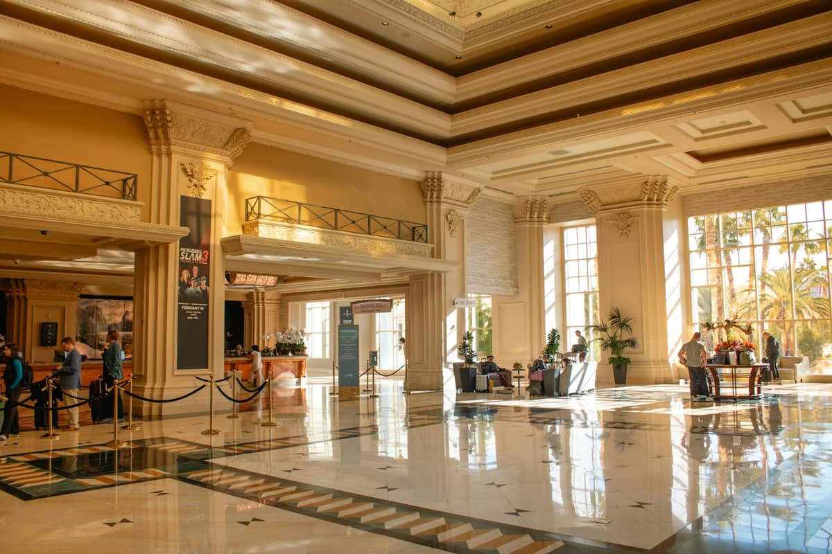 Spacious, sunlit hotel lobby in Las Vegas with people checking in and relaxing, marble floors, large windows, and palm trees visible outside.