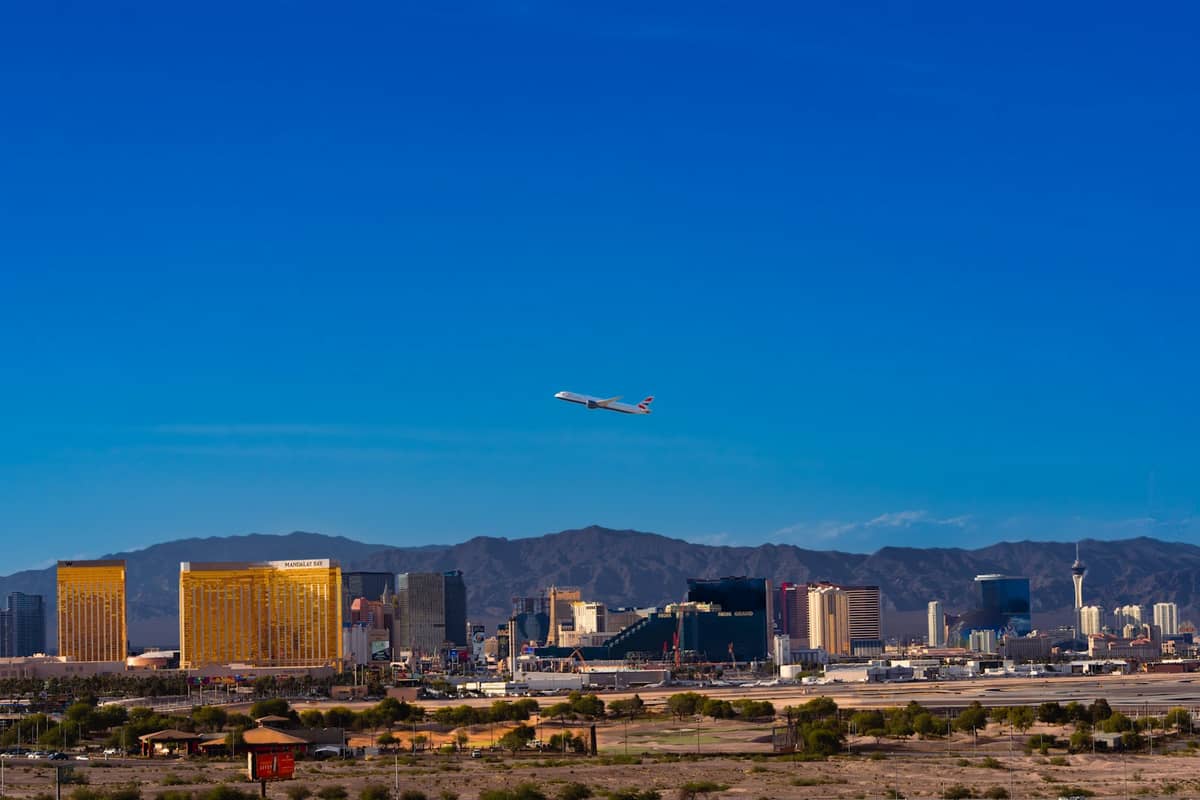 Wide view of Las Vegas Strip skyline under clear blue sky with distant desert mountains and an airplane taking off.