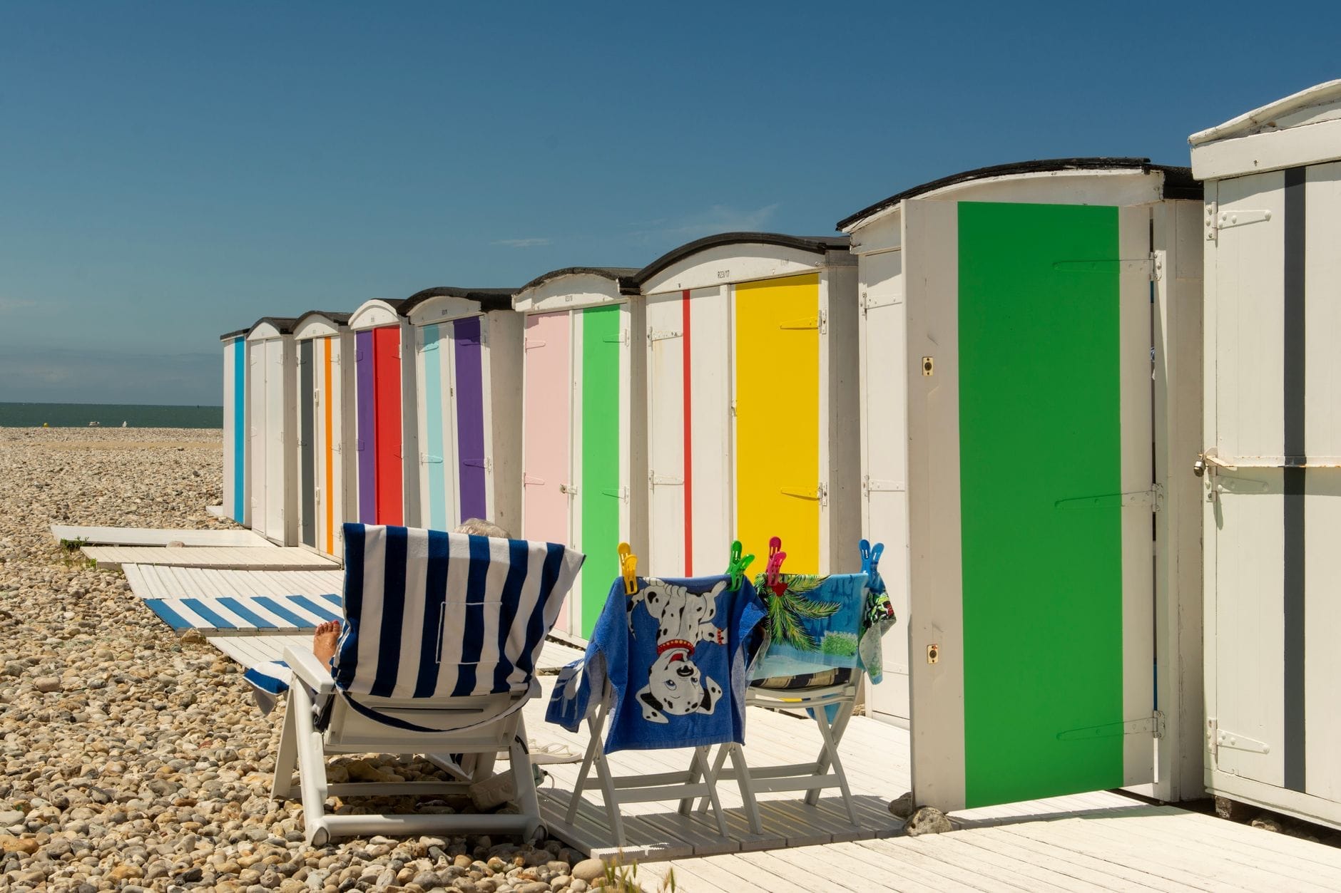 Colorful beach huts, striped lounge chairs, and summer clothes drying under a bright blue sky on Le Havre’s pebble beach.