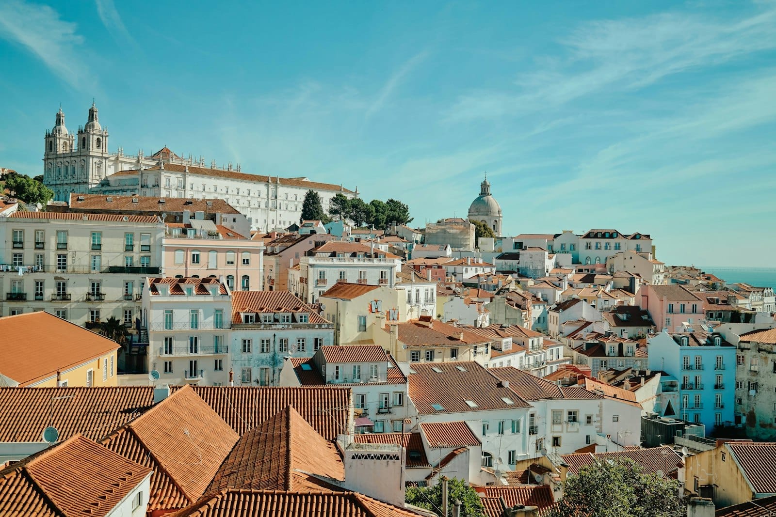Vista panorâmica do histórico bairro da Alfama em Lisboa, com prédios coloridos, telhados de cerâmica, torres icônicas e céu azul, transmitindo aquela atmosfera ensolarada e acolhedora da cidade.