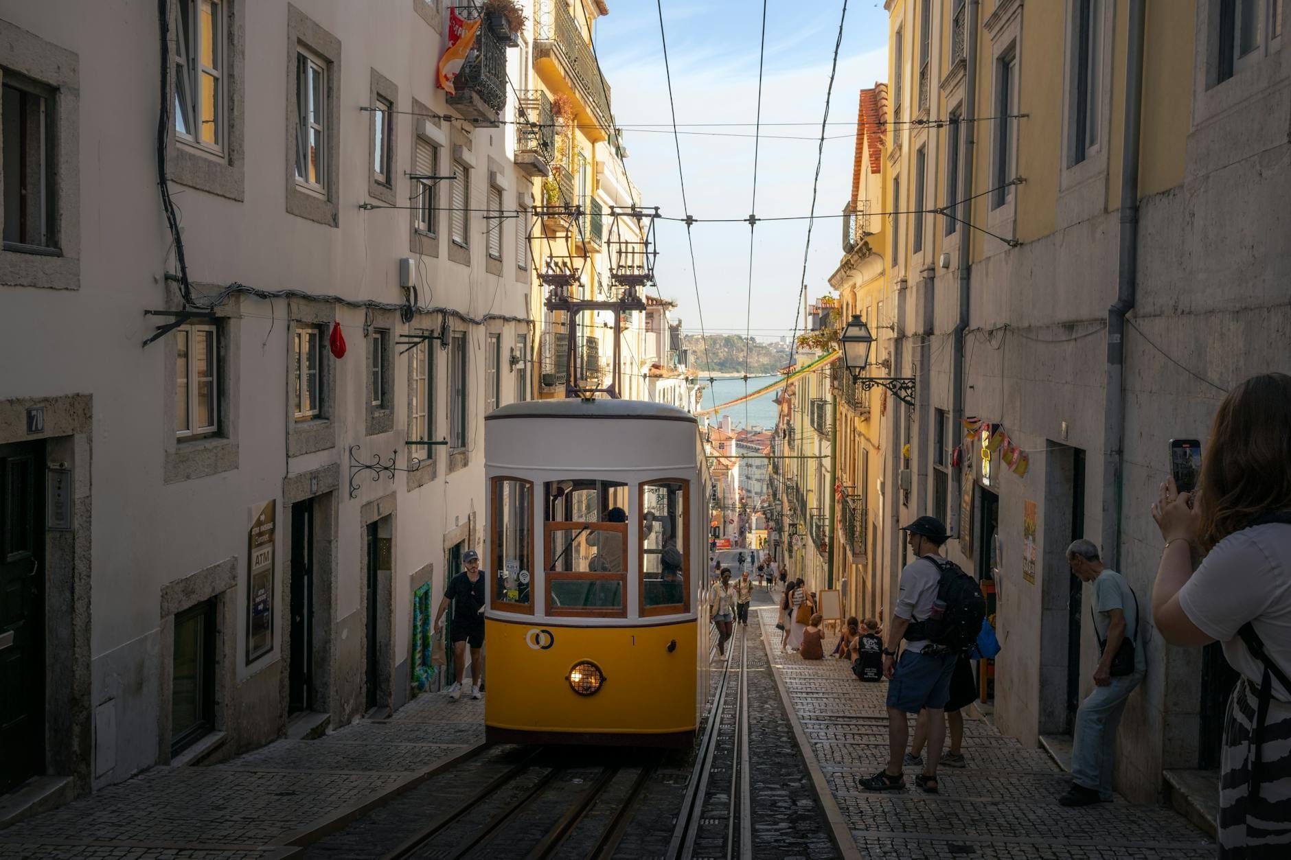 Rua íngreme de Lisboa com um eléctrico amarelo, pessoas em trajes de verão e luz solar intensa, captando a atmosfera animada de Lisboa no verão.