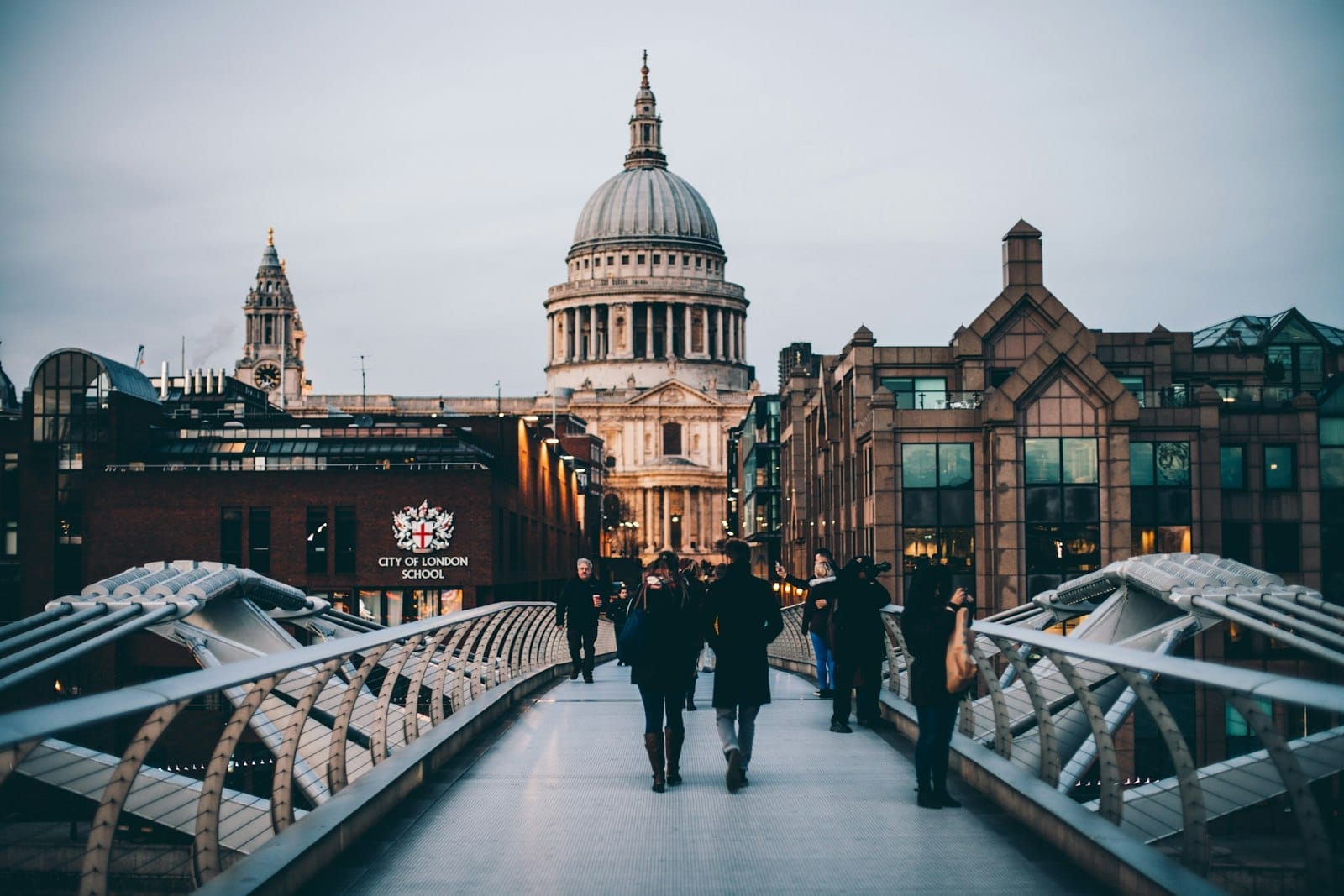 Vista da Catedral de St Paul's a partir da Millennium Bridge ao entardecer, com pessoas caminhando e edifícios da cidade em ambos os lados.
