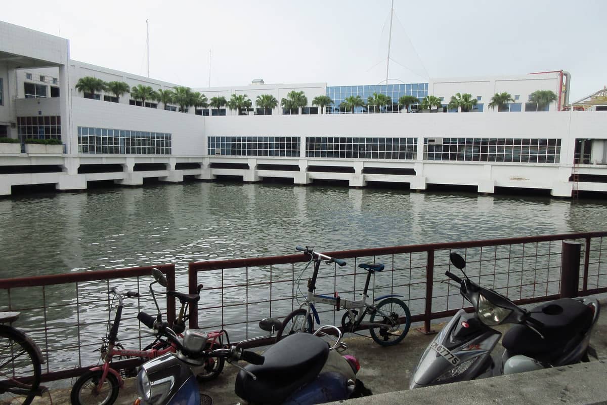 Das Äußere des Macau Outer Harbour Ferry Terminal mit Wasser davor und entlang eines Geländers geparkten Fahrrädern, an einem bewölkten Tag