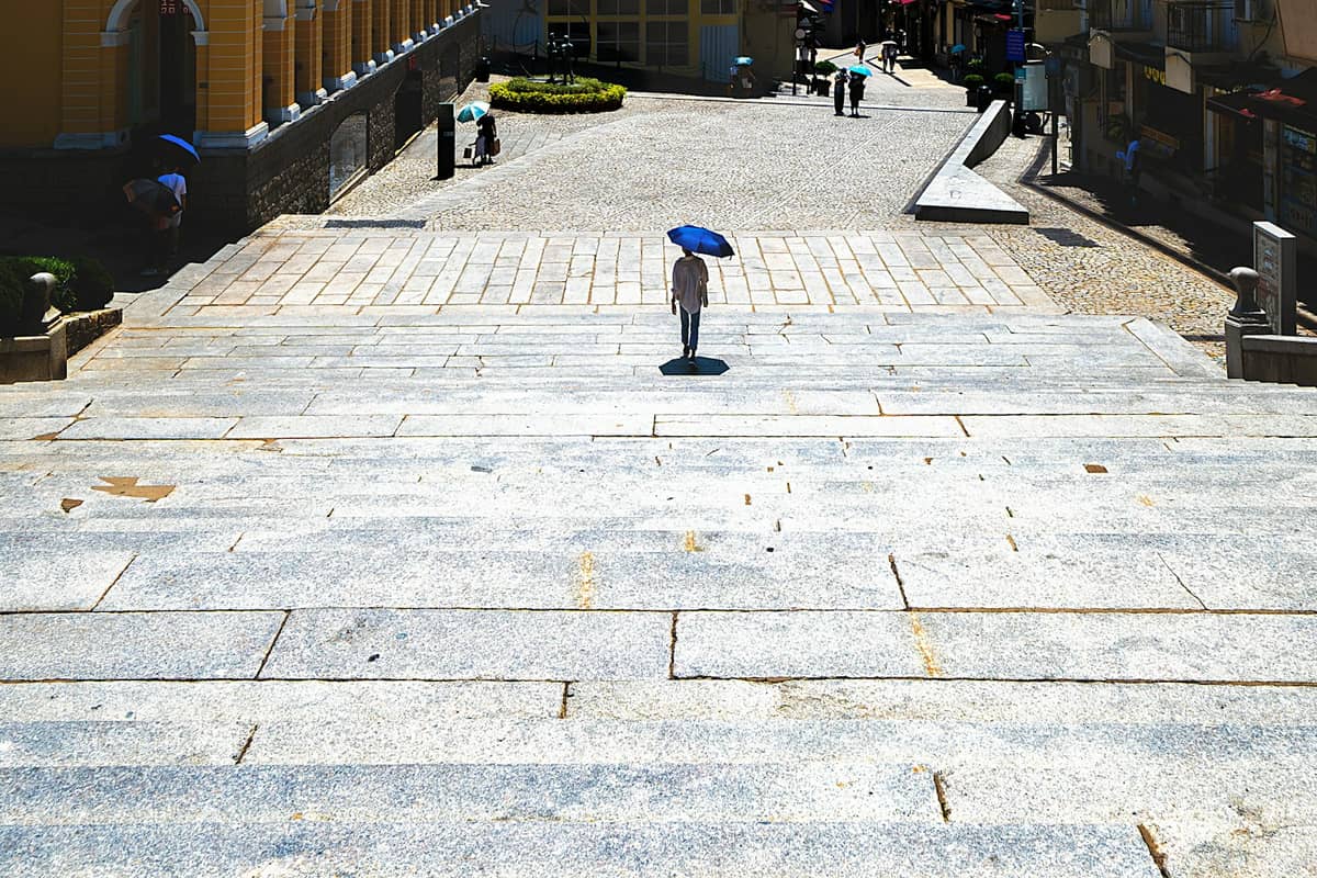 Ein fast leerer Steinplatz in Macau unter grellem Sonnenlicht, mit einer Person, die einen blauen Regenschirm als Sonnenschutz trägt.