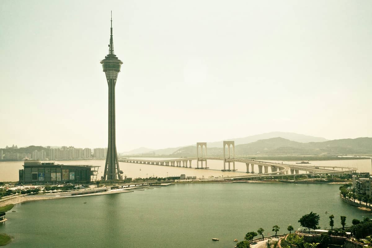 Weitwinkelpanorama des Macau Tower und einer großen Brücke mit Wasser im Vordergrund und der Skyline der Stadt im Hintergrund bei diesigem Tageslicht.