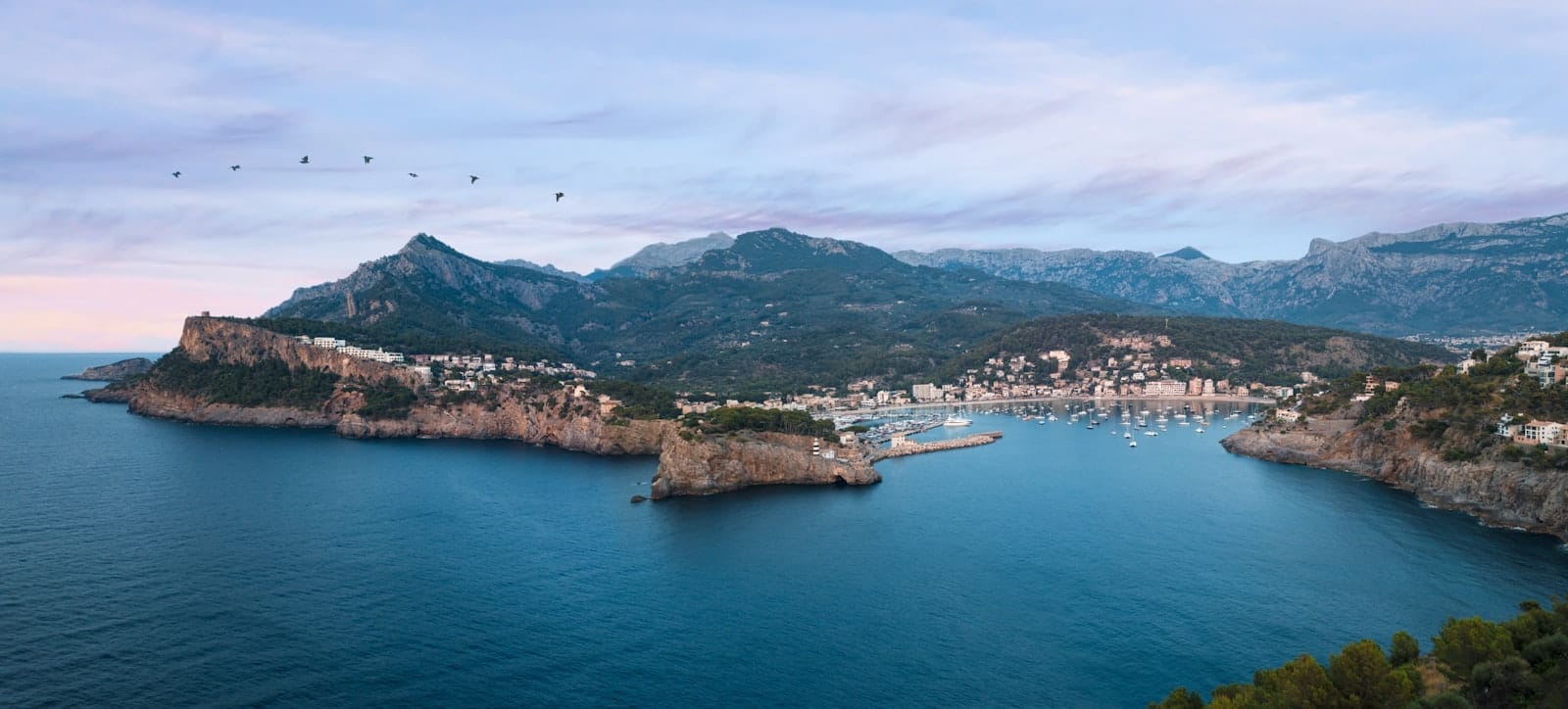 Vista panorâmica da costa de Mallorca com montanhas, uma marina repleta de barcos, mar azul e um céu sereno ao entardecer.