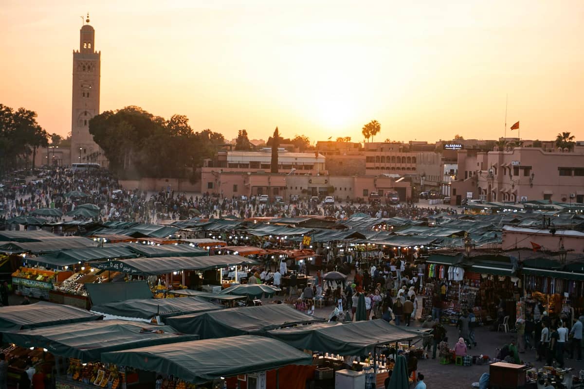 พระอาทิตย์ตกเหนือจัตุรัส Jemaa el-Fnaa ในเมือง Marrakech โดยมีมัสยิด Koutoubia เป็นฉากหลัง หลังคาตลาดสีเขียว และฝูงชนจำนวนมาก
