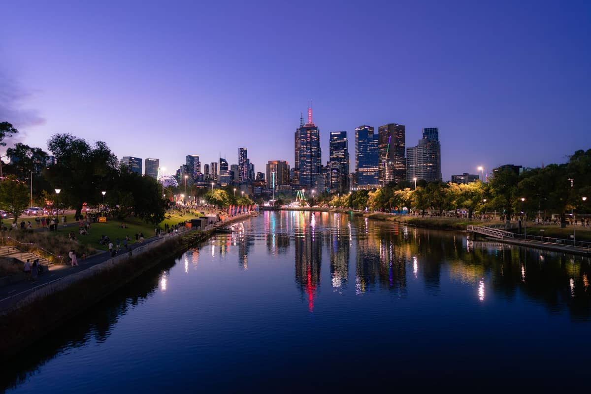 Horizonte de Melbourne al atardecer con rascacielos reflejados en el río Yarra y gente descansando a orillas del río.