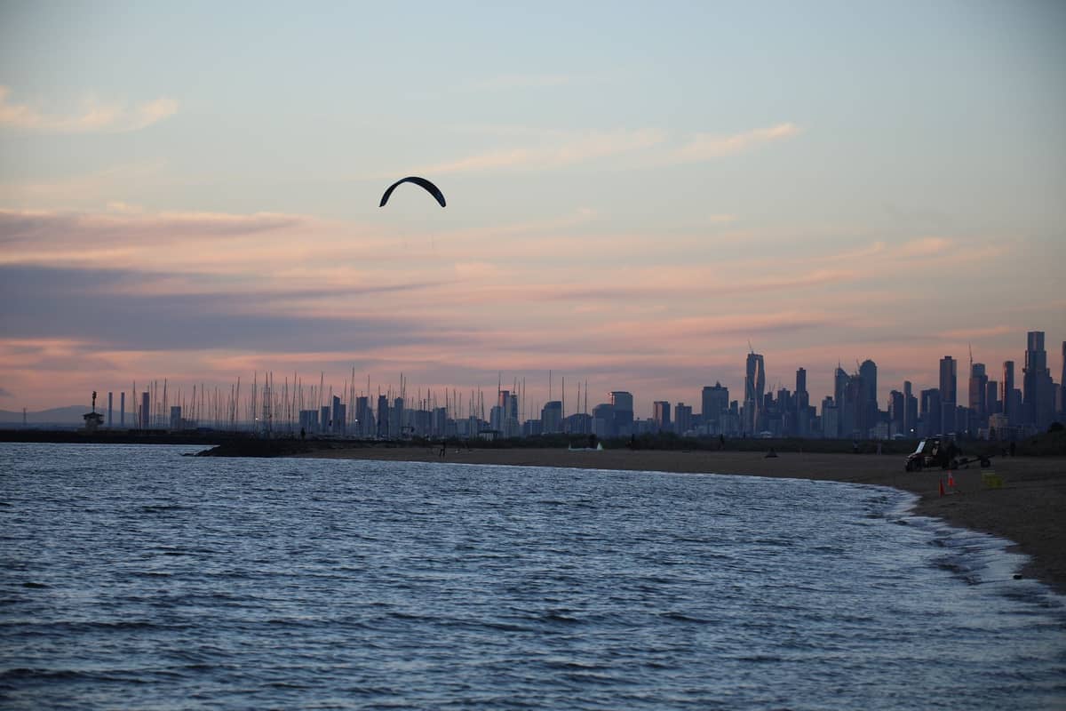 Skyline von Melbourne bei Sonnenuntergang, von einem Strand aus gesehen, mit Wasser, Booten und einem Kitesurfer am Himmel.