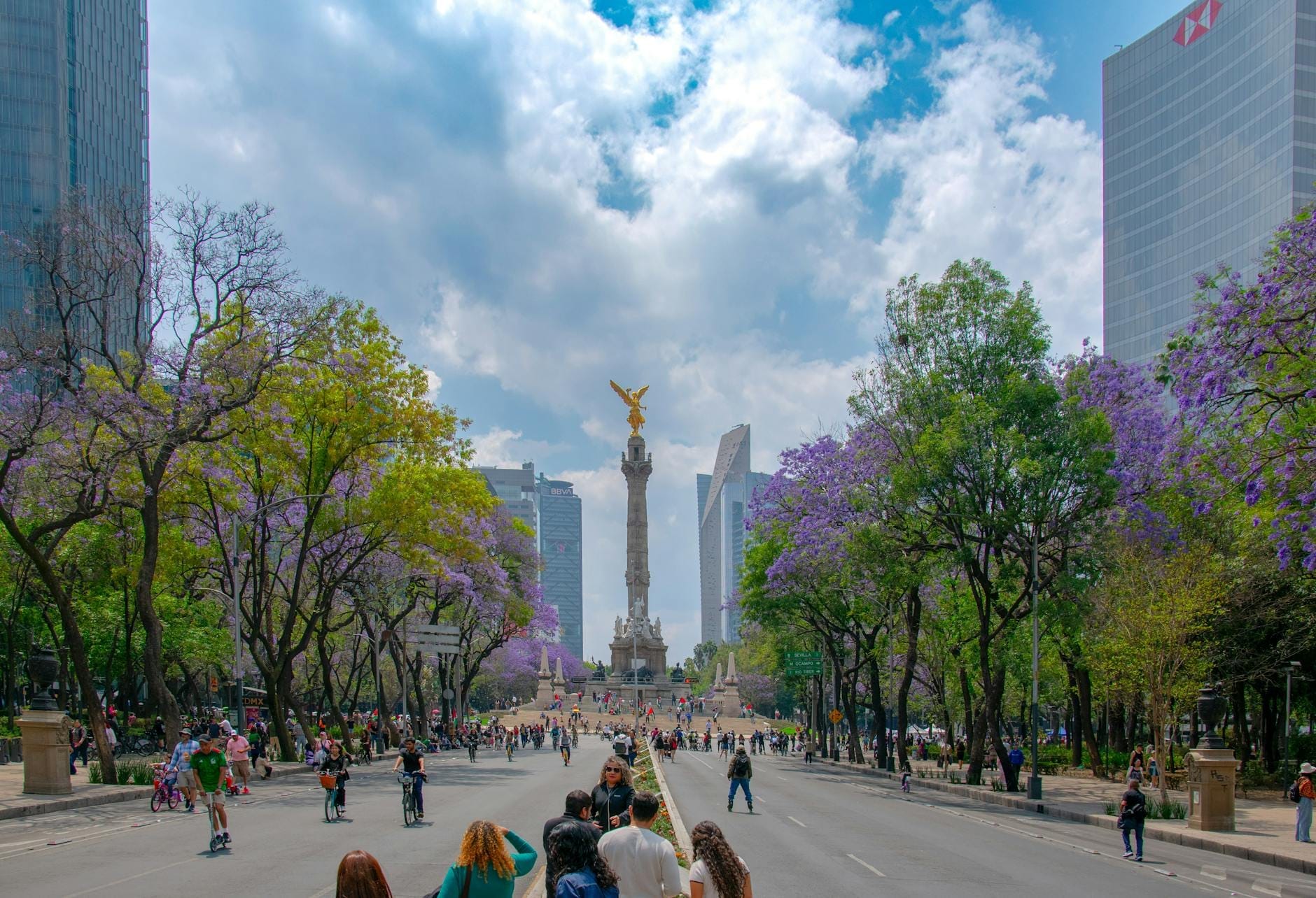 Vista do Ángel de la Independencia da Cidade do México com jacarandás em flor e pessoas aproveitando um dia ensolarado ao longo de uma grande avenida.