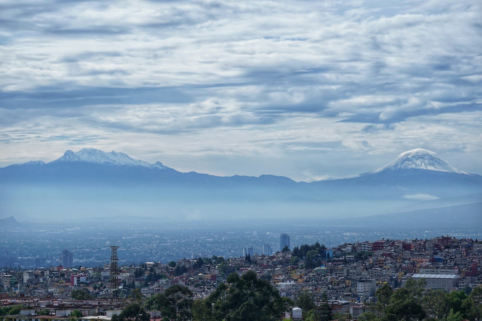 Vista ampla da Cidade do México com montanhas e vulcões ao fundo sob um céu parcialmente nublado, ilustrando o clima de alta altitude da cidade.