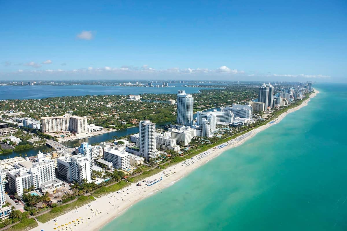 Aerial view of Miami Beach with turquoise water, white sand, and high-rise buildings under a clear blue sky.