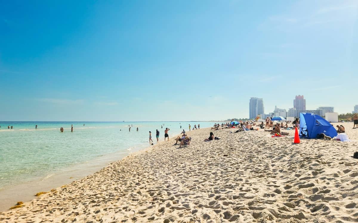 Sunny Miami Beach with people relaxing on sand and swimming, city skyline in background under clear blue sky.