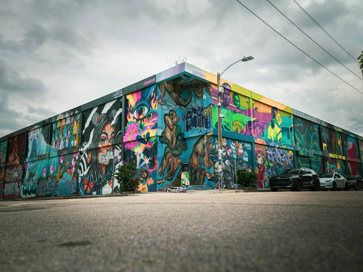 Vibrant Wynwood Walls building covered in colorful Miami street art murals under an overcast sky, with parked cars and empty street in foreground.