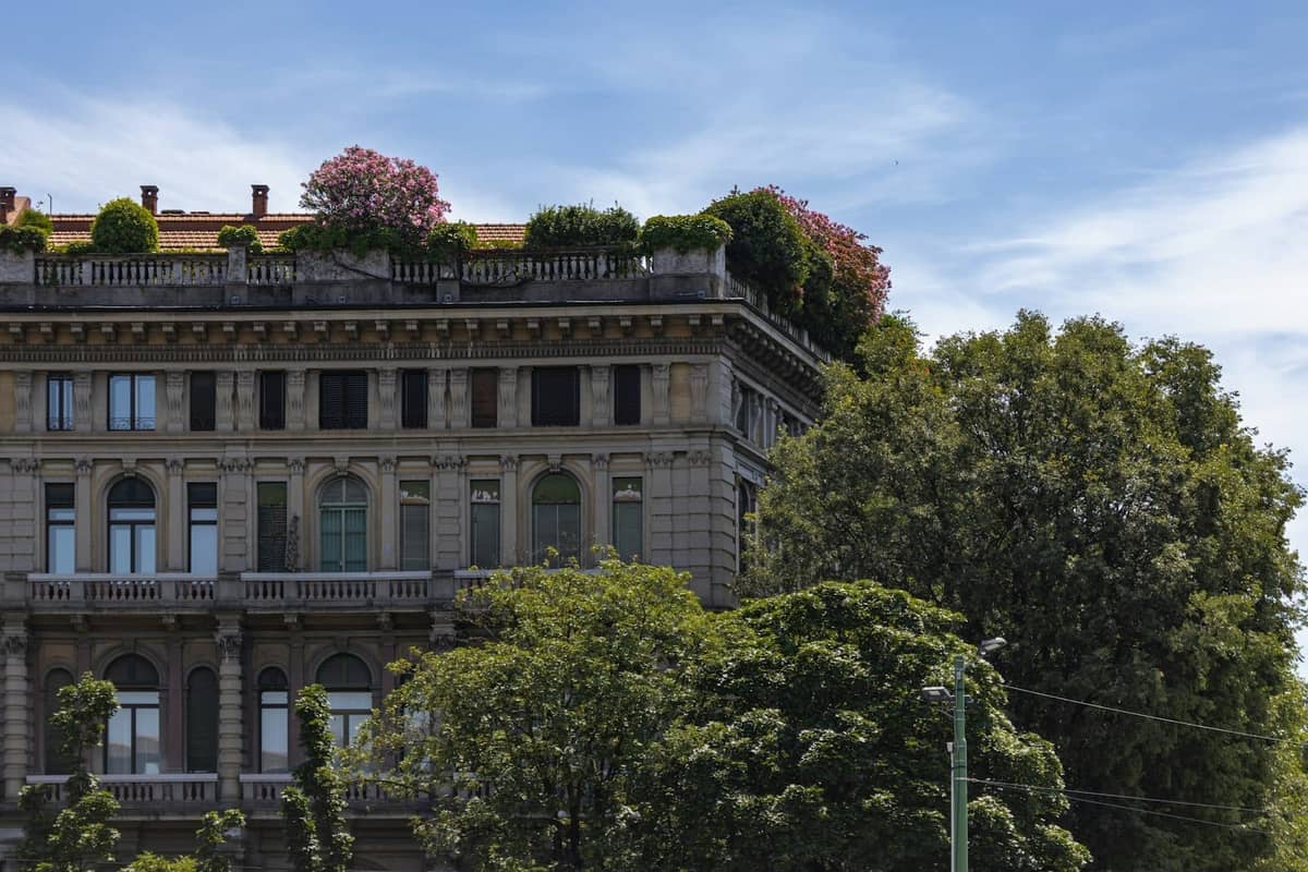 Elegante edificio storico a Milano con rigogliosi alberi verdi e fiori rosa in fiore sul tetto, sotto un cielo azzurro e limpido di primavera.