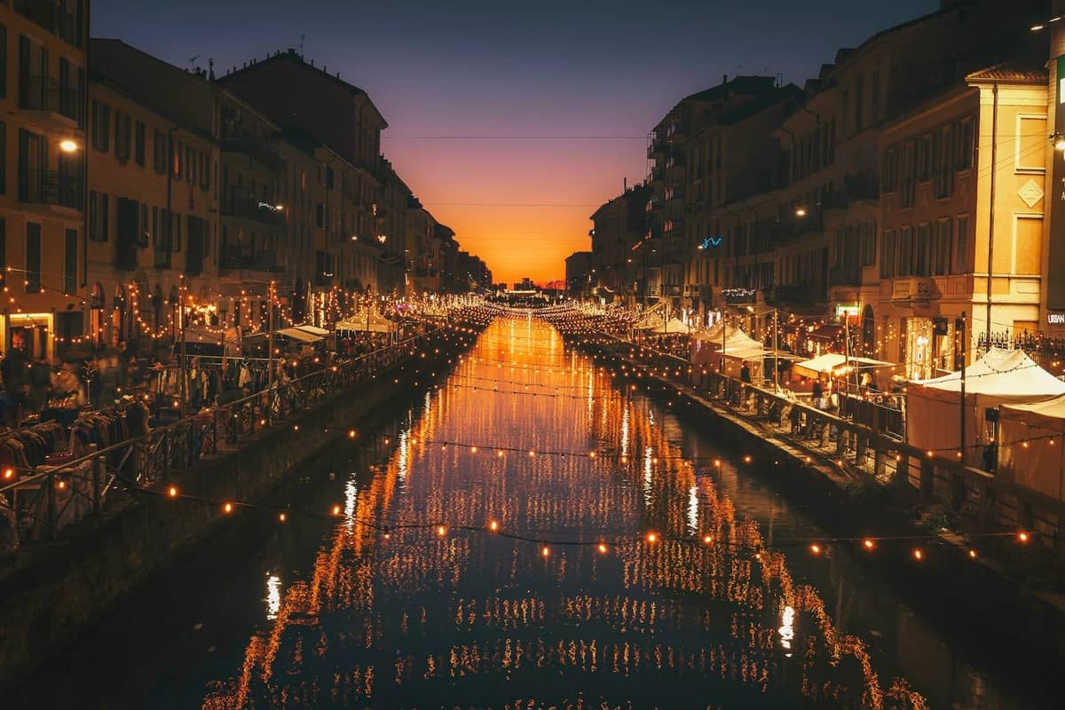 Il canale Navigli a Milano al tramonto, con luci festose, terrazze affollate e una vivace atmosfera estiva serale.