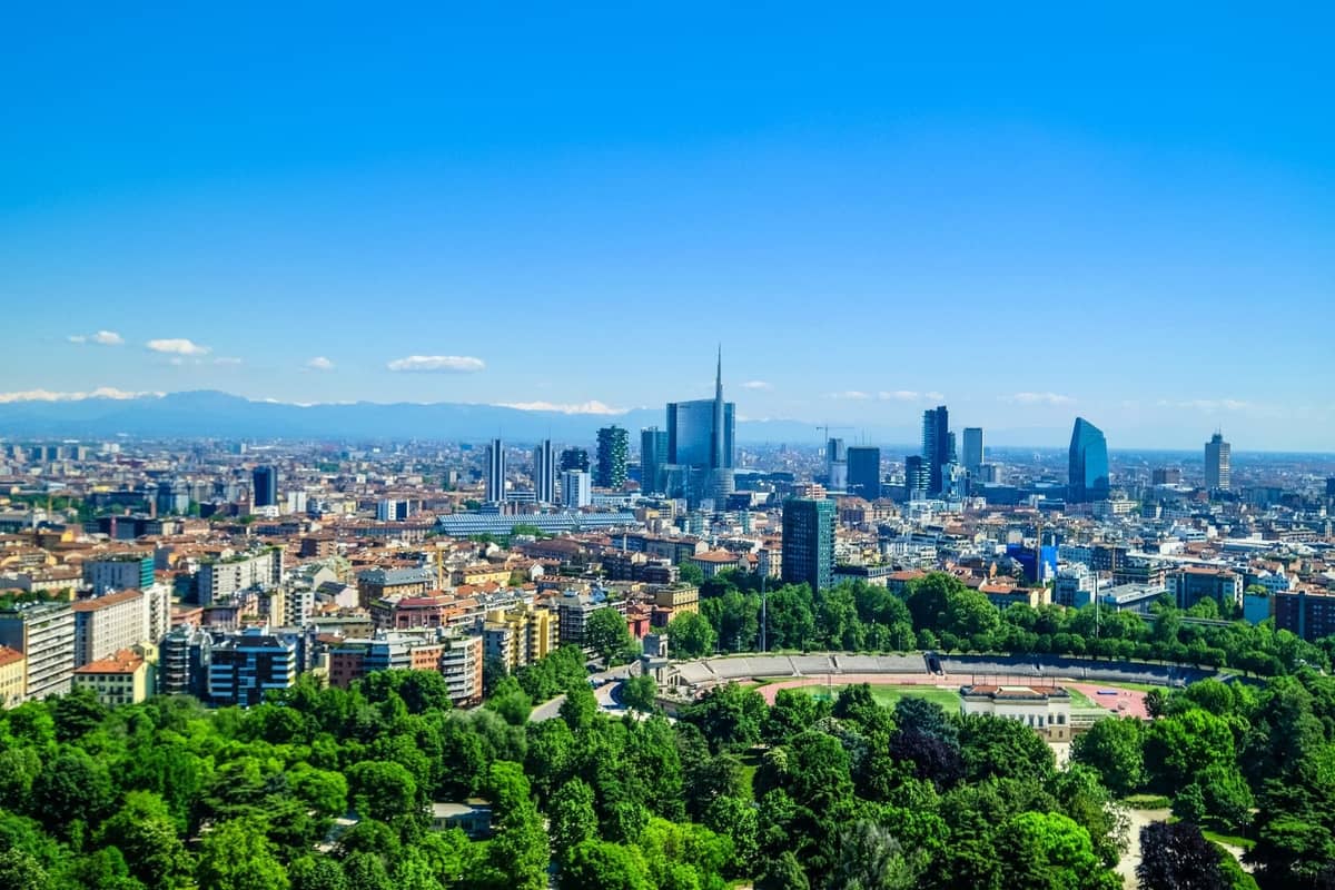Vista panoramica di Milano con gli edifici della città, parchi verdi e le Alpi visibili sullo sfondo sotto un cielo azzurro e soleggiato.