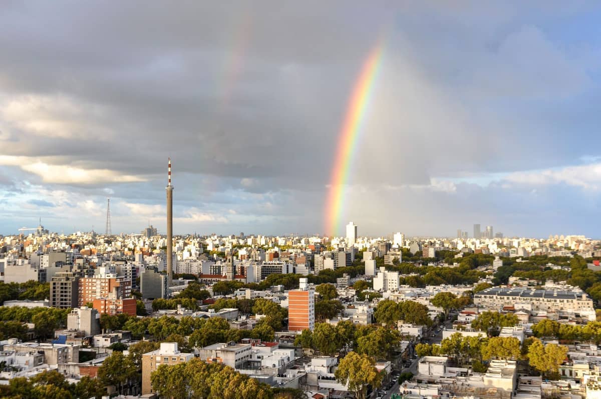 วิวพาโนรามาของทิวทัศน์เมือง Montevideo พร้อมอาคารสมัยใหม่ ต้นไม้สีเขียว และรุ้งกินน้ำพาดผ่านท้องฟ้าที่มีเมฆ