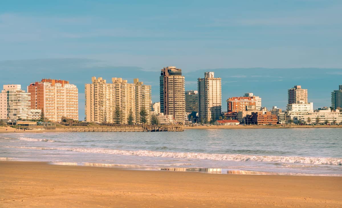 ชายหาดในเมืองที่มีทรายกว้างใหญ่ในมอนเตวิเดโอ พร้อมอาคารสูงสมัยใหม่ตลอดแนวริมน้ำ และคลื่นเบาๆ ใต้ท้องฟ้าแจ่มใส
