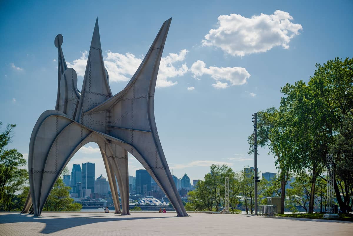 Große Außenskulptur mit der Skyline von Montreal im Hintergrund unter einem klaren, sonnigen Sommerhimmel, von Bäumen eingerahmt.