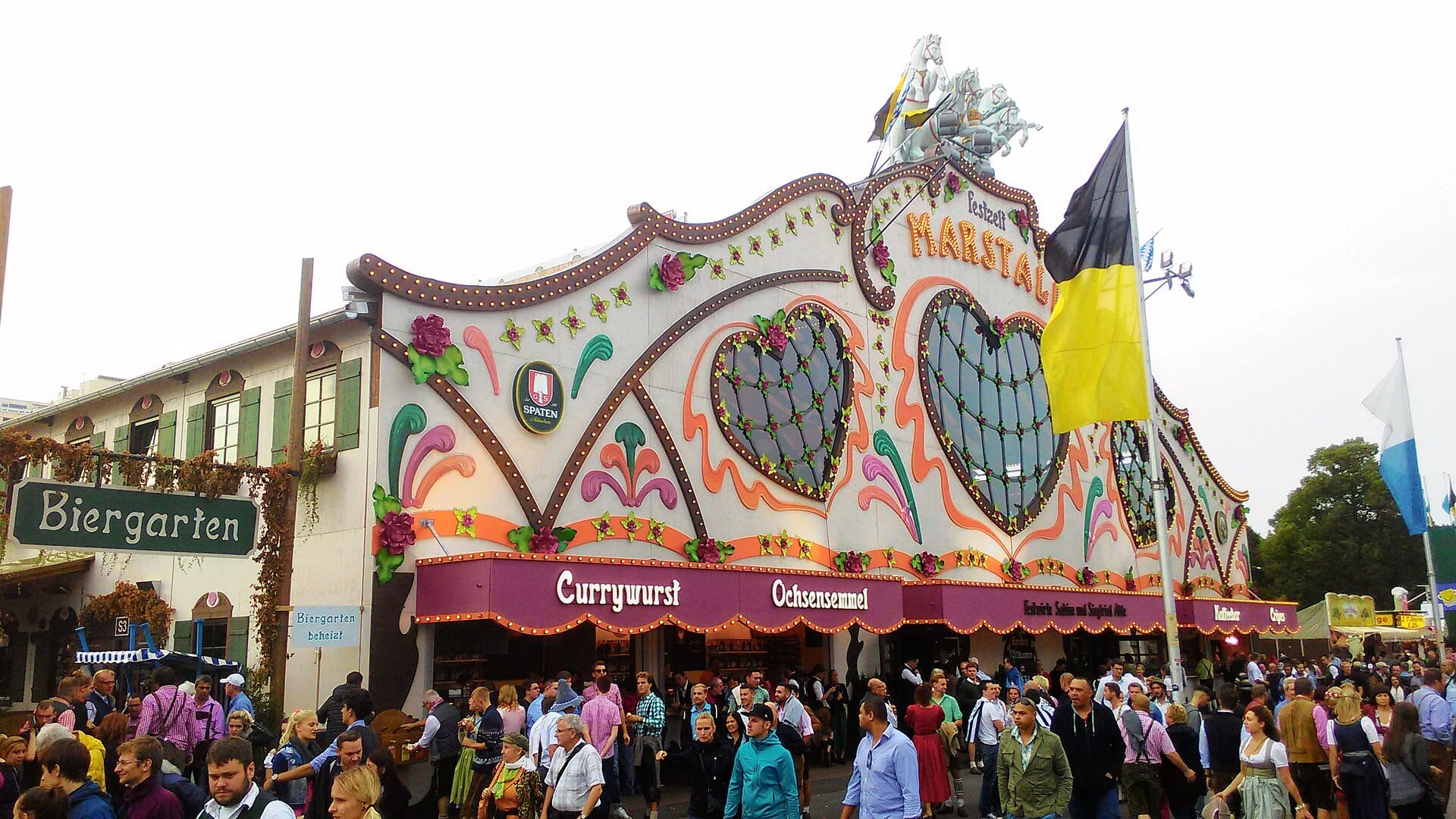 เต็นท์ Oktoberfest ขนาดใหญ่ตกแต่งด้วยรูปหัวใจและดอกไม้ ฝูงชนสวมชุดพื้นเมืองดั้งเดิม และป้าย Biergarten ใน München ช่วงเทศกาล