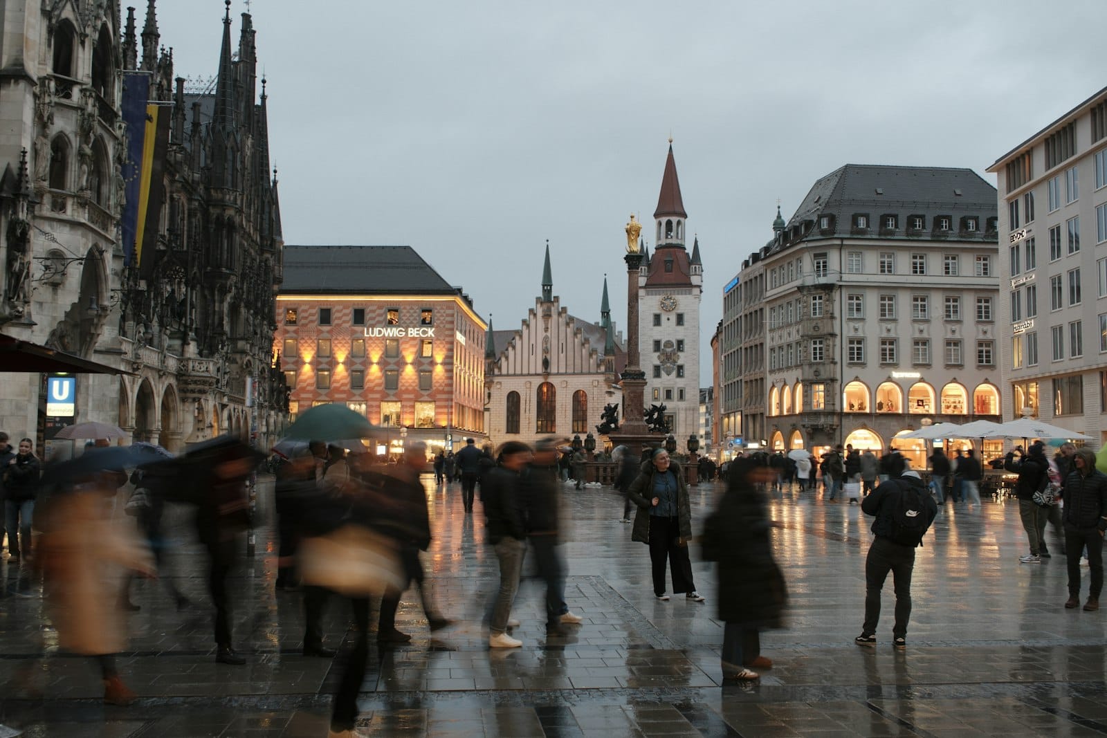 วันที่ฝนตกและท้องฟ้ามืดครึ้มที่ Marienplatz ในมิวนิก พร้อมผู้คนกลุ่มเล็กๆ และอาคารประวัติศาสตร์ สะท้อนบรรยากาศเงียบสงบนอกฤดูกาลท่องเที่ยว