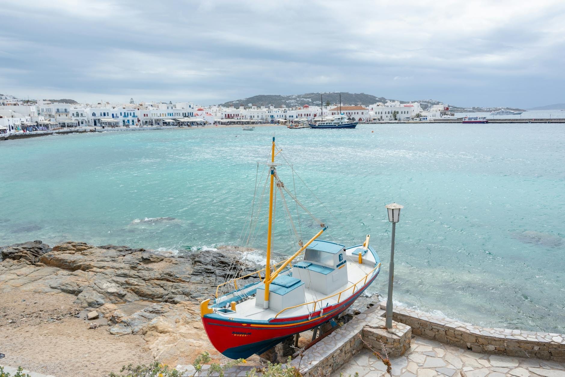 Un barco de pesca varado en un tranquilo frente marítimo nublado con el pueblo de Mykonos y la orilla desierta al fondo