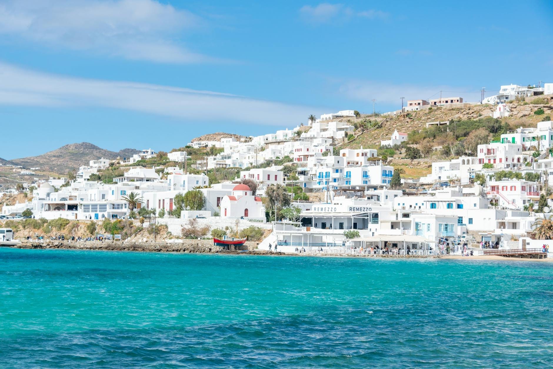 Paseo marítimo de Mykonos iluminado con edificios encalados en una colina y mar turquesa bajo cielo azul, ilustrando el clima mediterráneo.