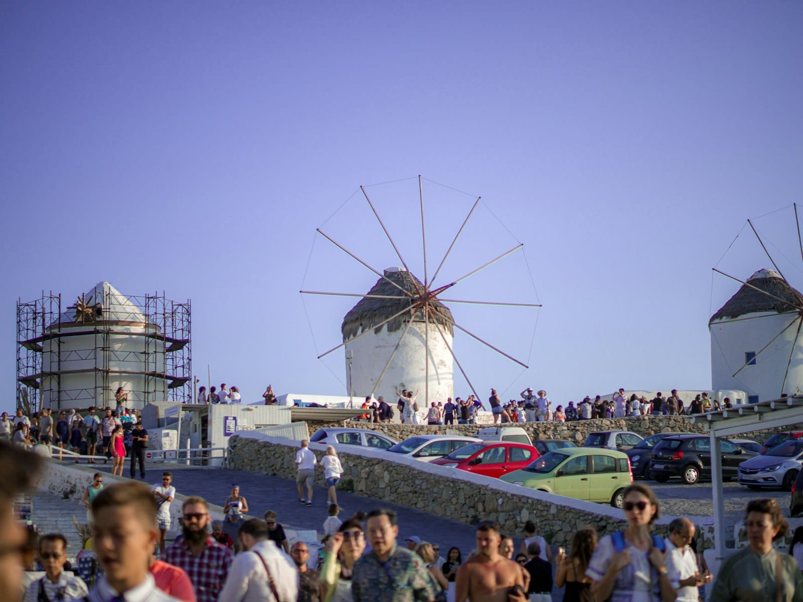 Escena concurrida en los molinos de Mykonos con muchos turistas y coches bajo un cielo despejado, ilustrando la afluencia de visitantes en temporada alta.