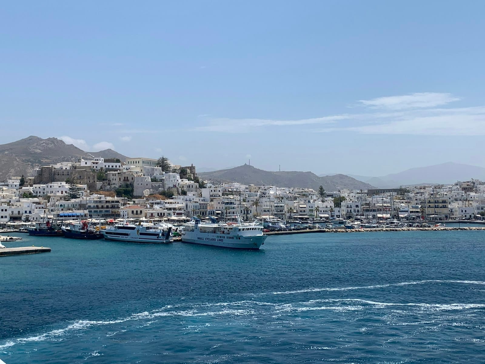 Ferrys y barcos atracados en el puerto de Mykonos con edificios blancos de la ciudad al fondo bajo un cielo despejado.