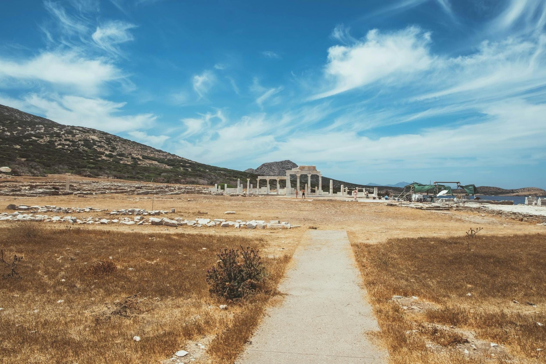 Camino que conduce a ruinas antiguas bajo un cielo azul, con columnas y colinas al fondo en Mykonos.