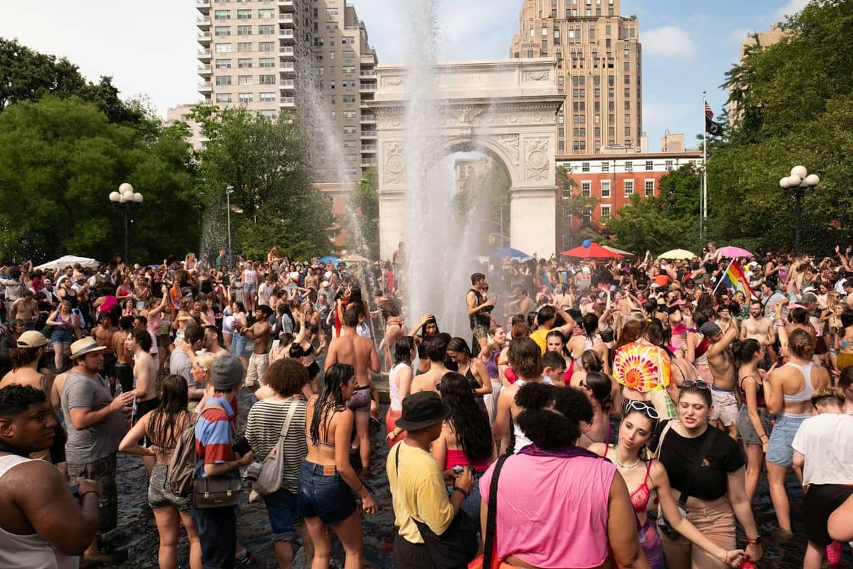 Eine große Menschenmenge genießt einen Sommertag rund um den Brunnen im Washington Square Park mit dem Bogen und der Stadtsilhouette im Hintergrund.
