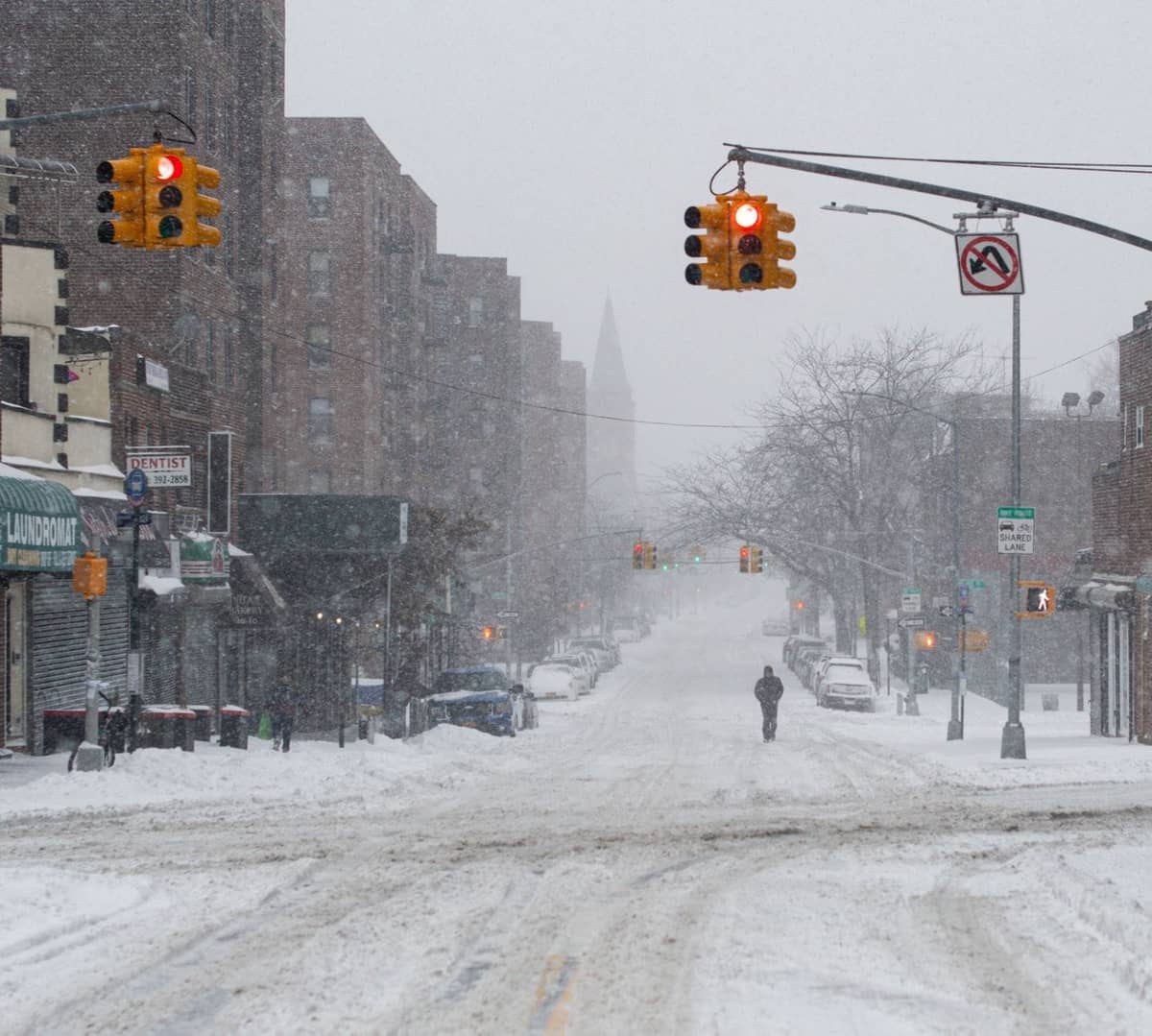 Schneebedeckte Straße in New York City mit Ampeln, Gebäuden und einer einzelnen Person, die während eines heftigen Winterschneefalls spaziert.