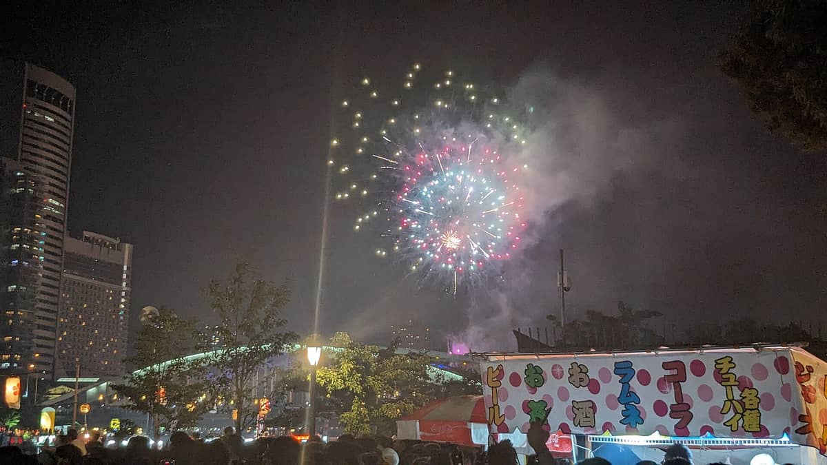 Colorful fireworks light up the night sky above Osaka city, with crowds and festival food stalls in the foreground.