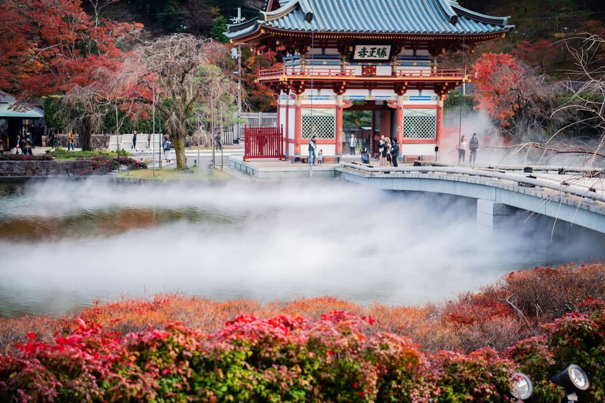 Traditional Japanese gate and bridge surrounded by vibrant autumn foliage with mist rising from a pond in Osaka.