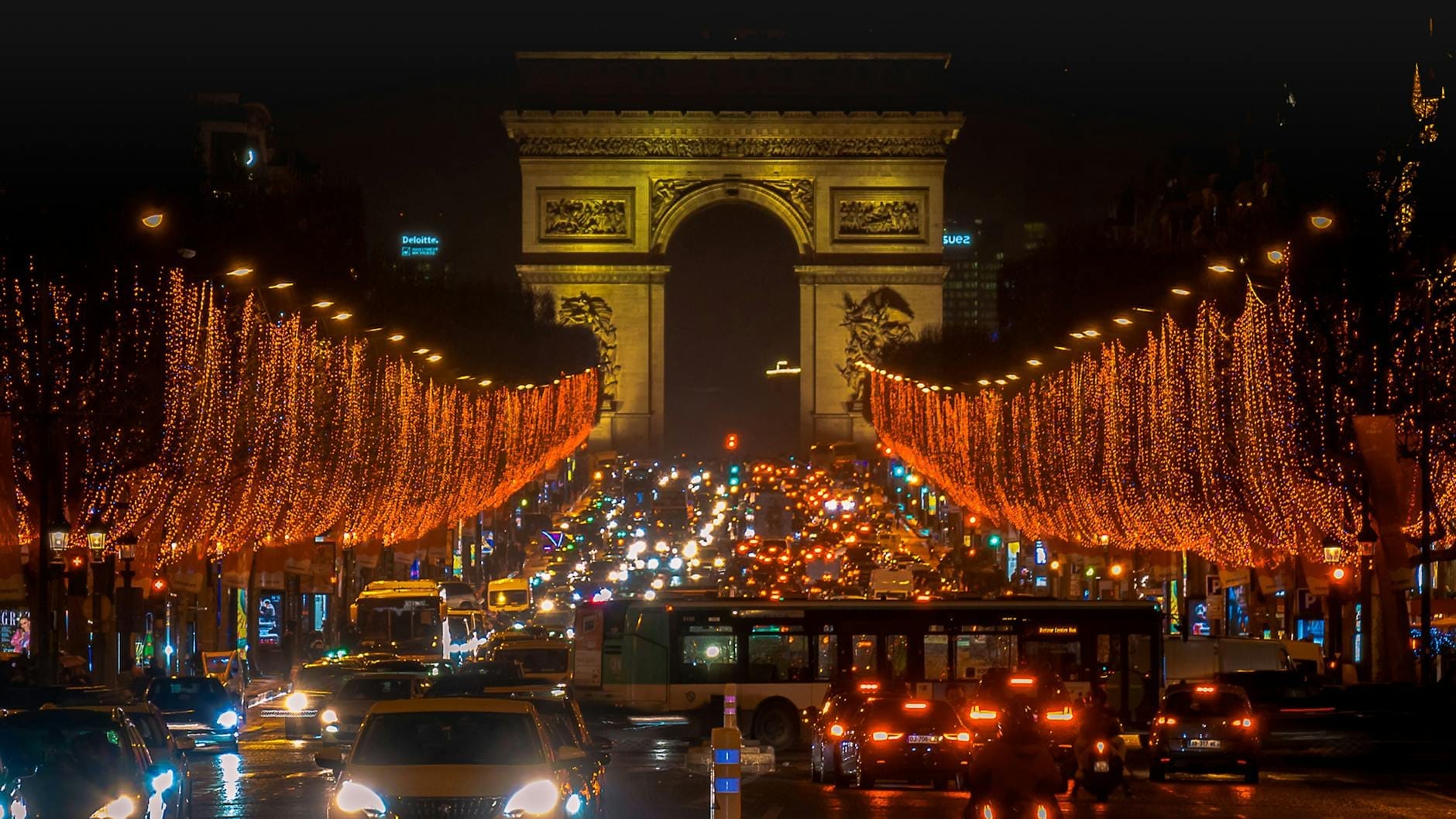 Blick auf den Arc de Triomphe bei Nacht mit festlichen Feiertagslichtern entlang der Champs-Élysées und lebhaftem Verkehr darunter.