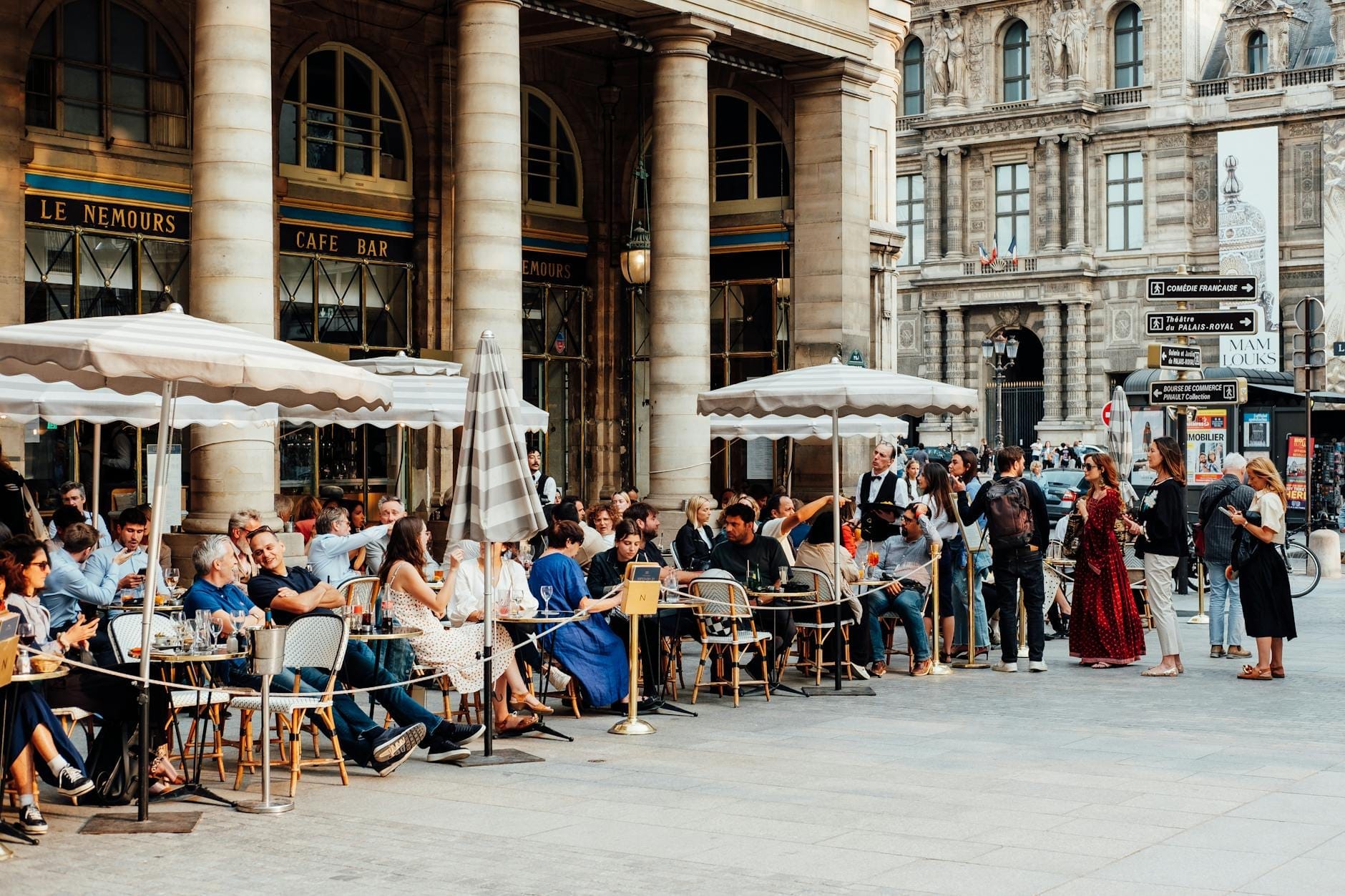 Belebte Pariser Außencafé-Szene mit Menschenmassen, die an sonnigen Tagen unter Sonnenschirmen in der Nähe historischer Gebäude speisen und sich unterhalten.